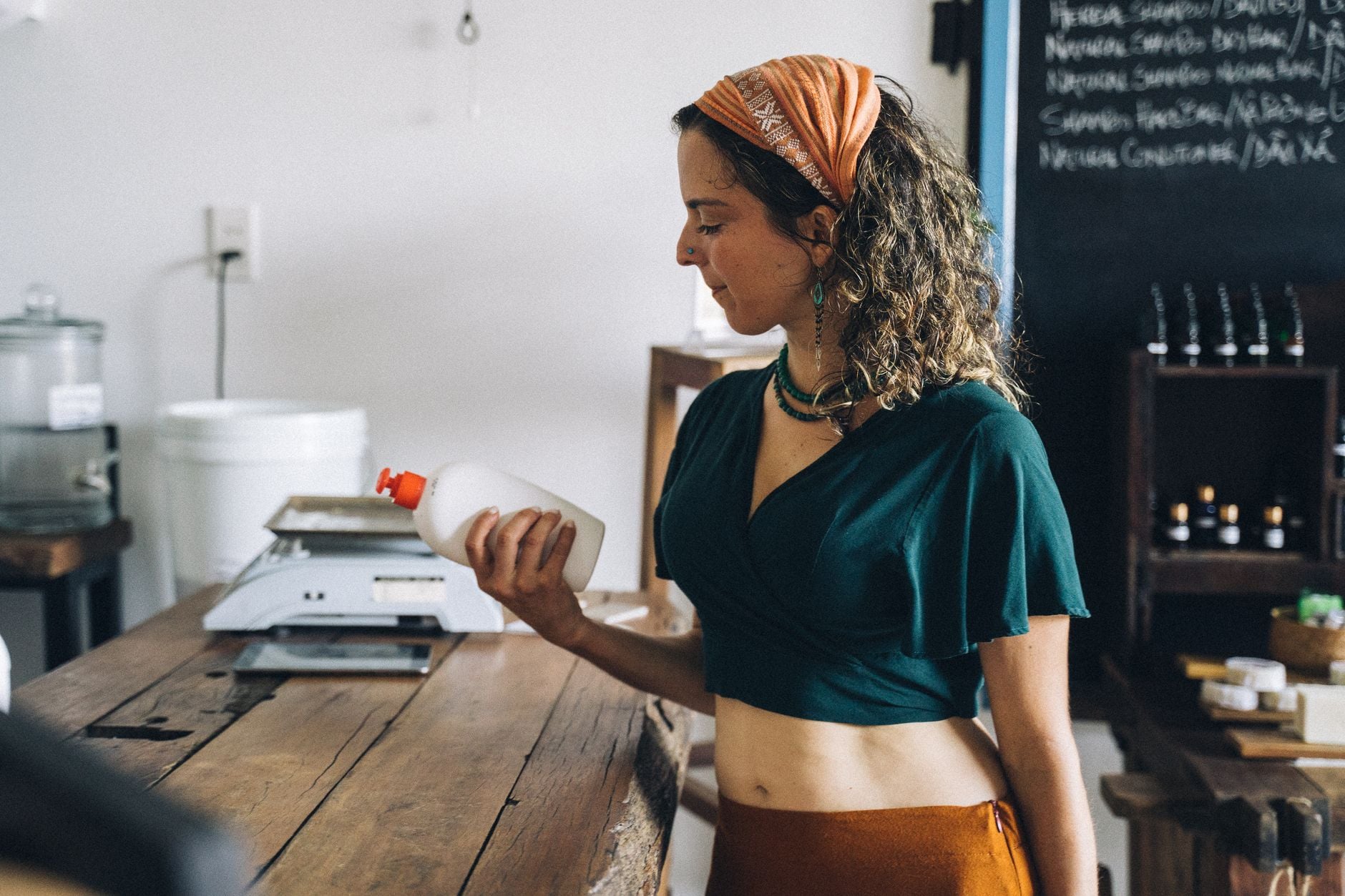 Woman in teal shirt at wooden table examining white product bottle in beauty product workshop