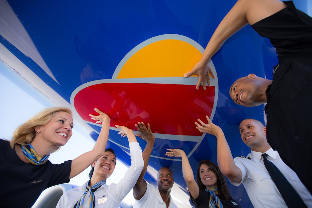Diverse airline employees in uniforms smiling while holding red and yellow heart-shaped prop in corridor